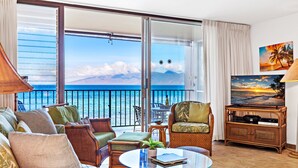 A living room with a large sliding glass door leading to a balcony with a view of the ocean. There is a television on a wooden stand and a small table with a plant. - Oceanfront Maui Escape with Private Lanai, Pool & Stunning Views (Lahaina)