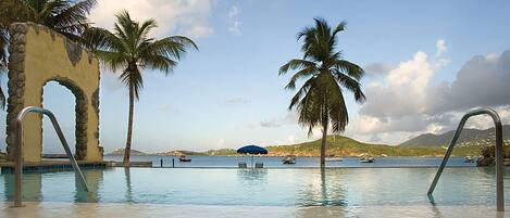 A swimming pool with palm trees, a stone archway, and a view of boats on the water.