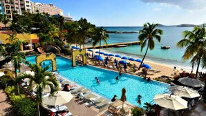 A resort pool area with a view of the ocean, palm trees, and a hotel building in the background.