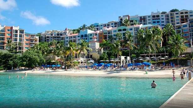A beachfront hotel with multiple buildings, palm trees, and a sandy beach with umbrellas.