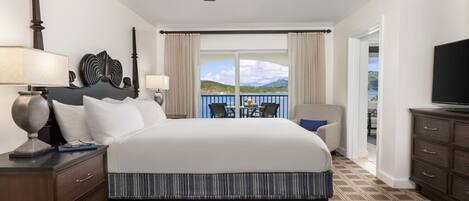 A hotel room with a large bed, a ceiling fan, a TV, a balcony with a view of the landscape, and a dining area with chairs and a table.