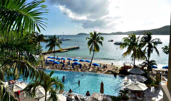 A pool area with umbrellas, palm trees, and a view of the sea with boats.