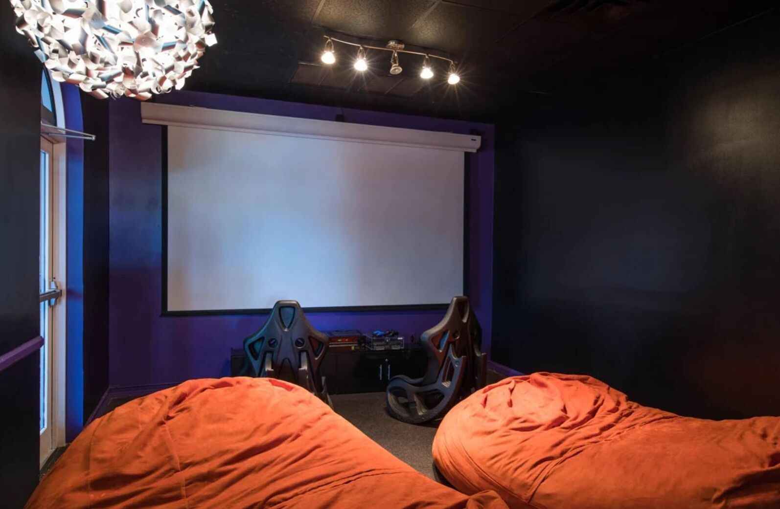 A room with a large screen, two gaming chairs, and orange bean bags.