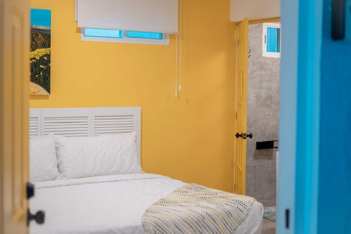 A bedroom with a bed, a window with blinds, a door, and a wall with a painting.