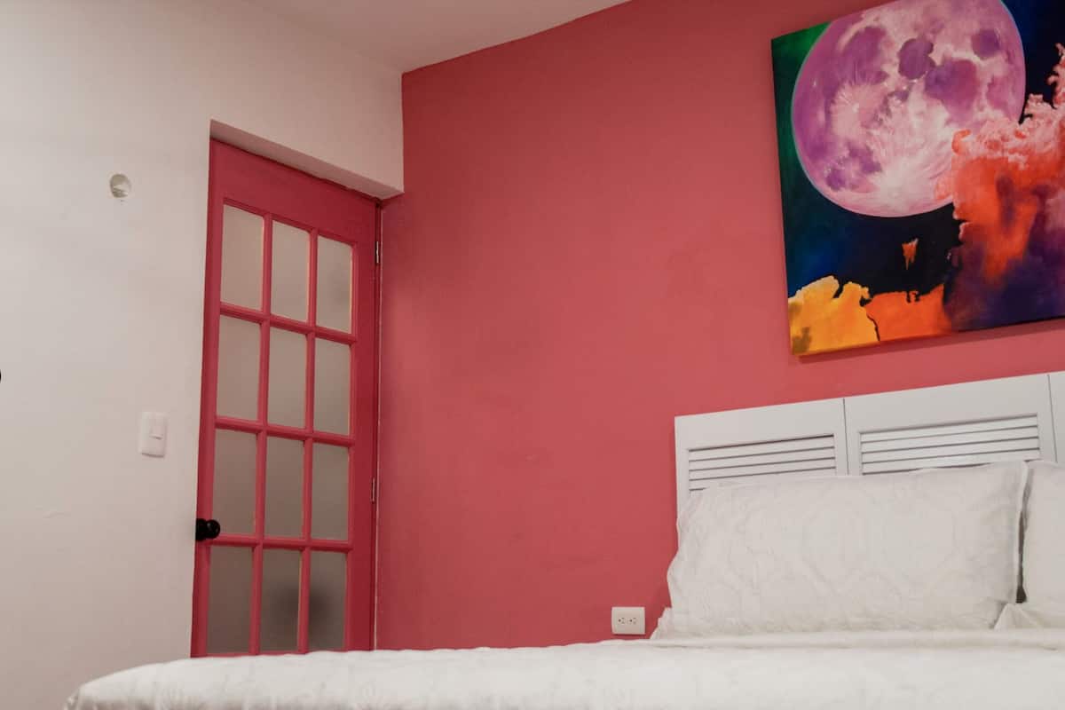 A bedroom with a pink wall, a white bed, and a painting of a moon.