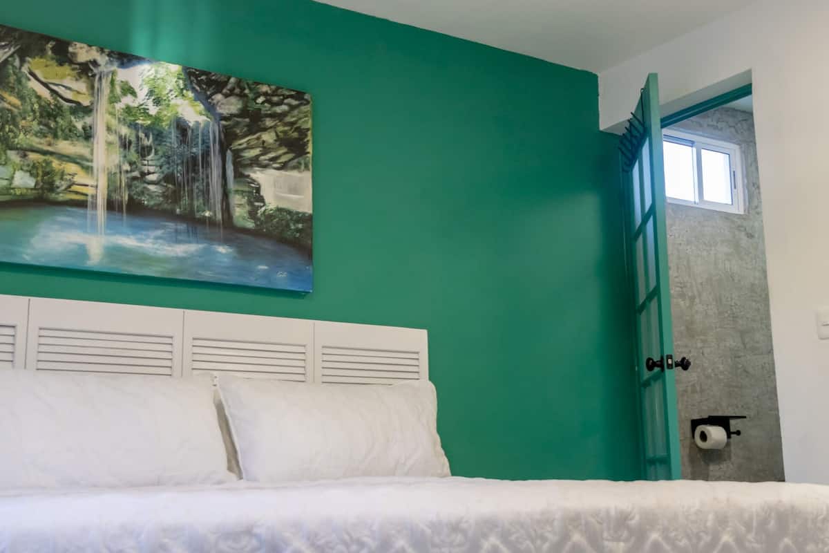 A bedroom with a bed, a green wall, a painting of a waterfall, and a door with a window.