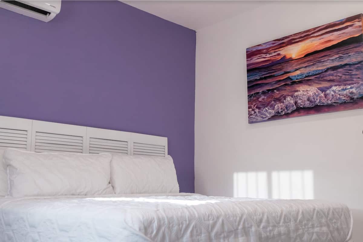 A bedroom with a bed, white headboard, and a large painting of a sunset over the ocean.