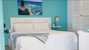 Une chambre avec un lit blanc, des murs de couleur turquoise, un tableau représentant une sirène et une fenêtre avec des stores.