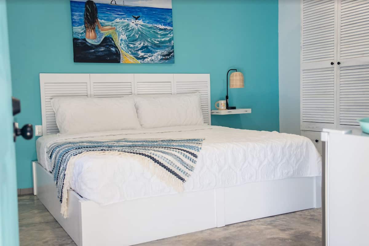 A bedroom with a white bed, teal walls, a painting of a mermaid, and a window with blinds.