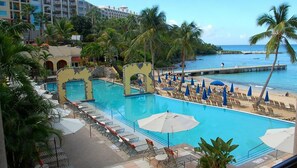 Ein Poolbereich mit Liegestühlen und Sonnenschirmen, eine Pier, die ins Wasser führt, und im Hintergrund Palmen sowie ein Hotelgebäude.