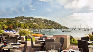 An outdoor dining area with tables and chairs, overlooking a marina with boats and a hillside with houses.