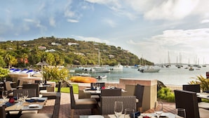 An outdoor dining area with tables and chairs, overlooking a marina with boats and a hillside with houses.