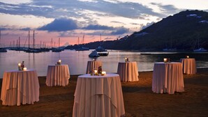 A beachside setting with multiple round tables set up for dining, overlooking a calm body of water with boats and a hilly landscape in the background.