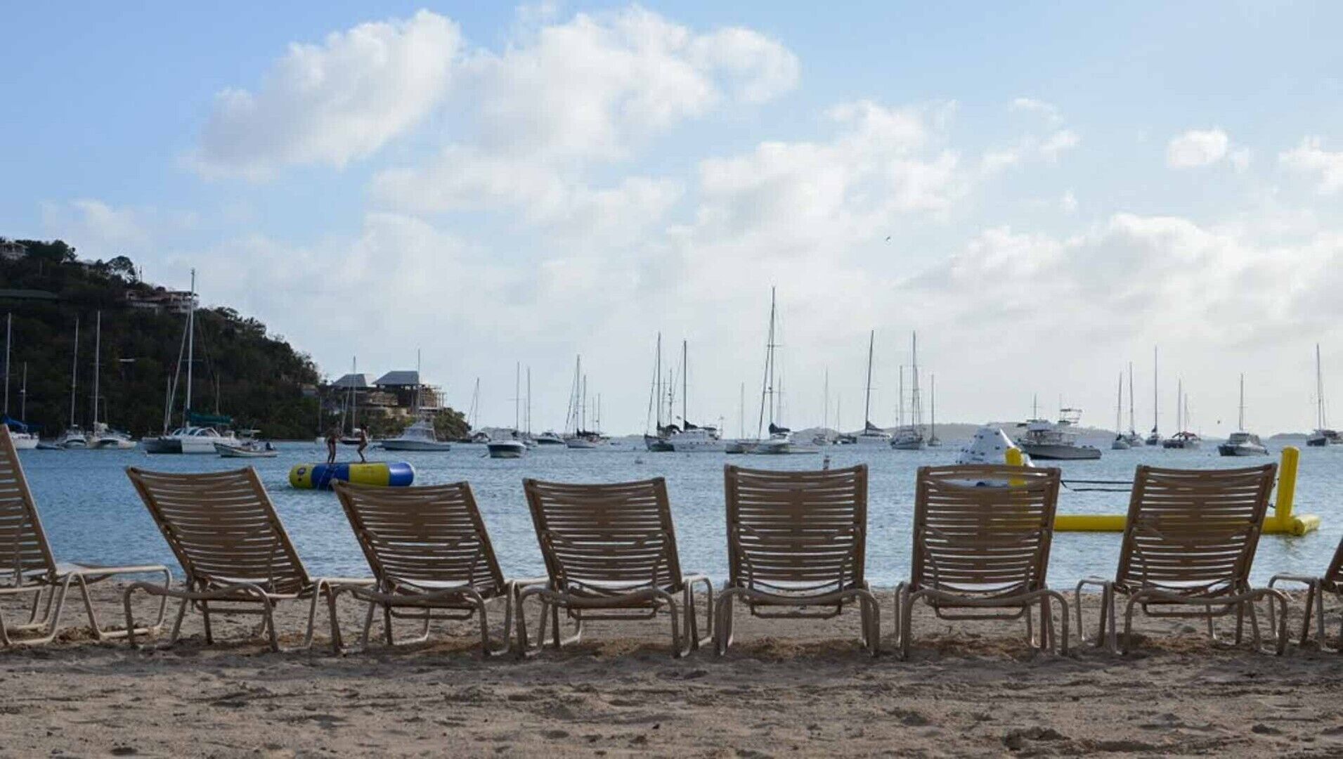 A beach with rows of wooden chairs facing the sea, a sandy shore, and several sailboats in the background.