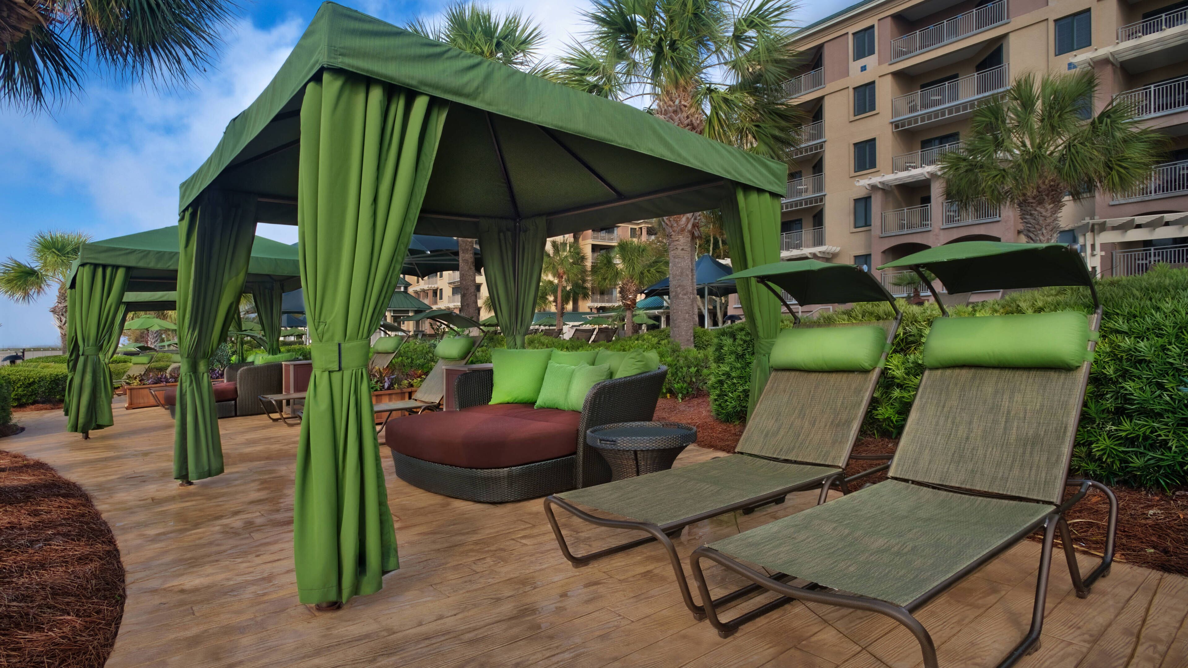 Un área de estar al aire libre con toldos verdes, sillones reclinables acolchados y una terraza de madera.
