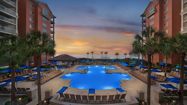 A resort pool area with lounge chairs and umbrellas, surrounded by palm trees and overlooking the ocean at sunset.