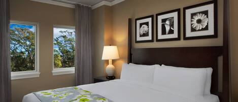 Ein Hotelzimmer mit einem großen Bett, einem Nachttisch mit Lampe, drei gerahmten Bildern an der Wand und einem Fenster mit Vorhängen.