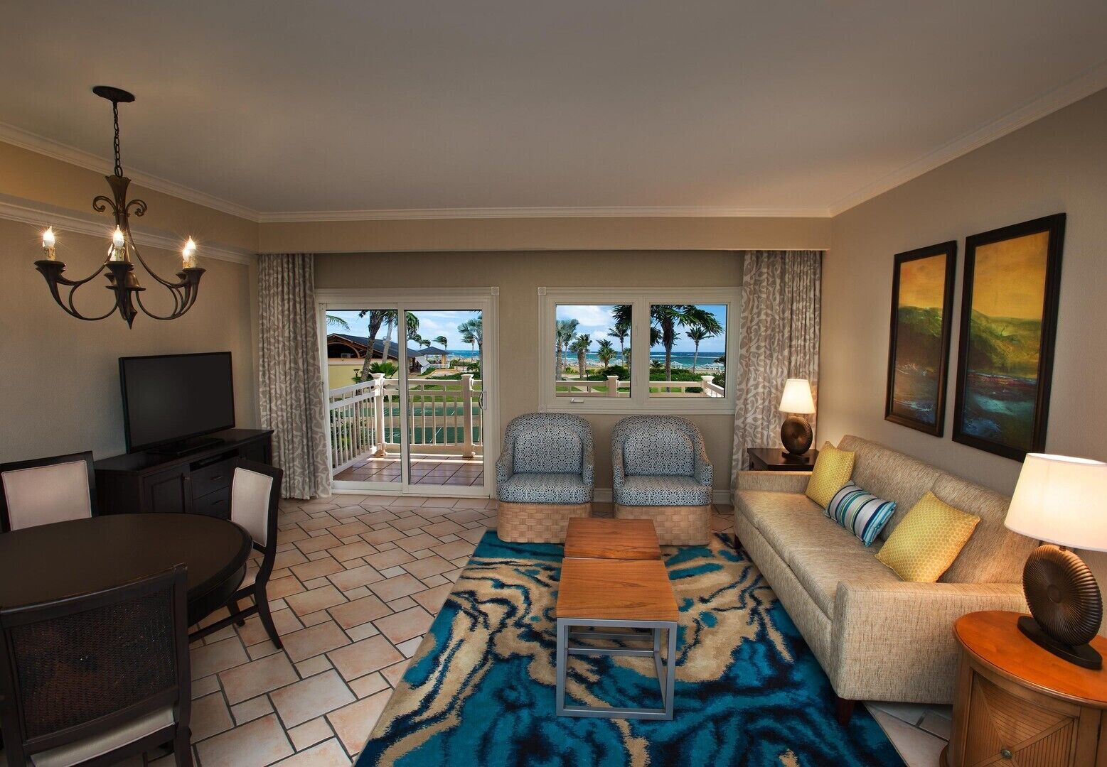A living room with a TV, dining table, chairs, a sofa, and a view of the beach through large windows.