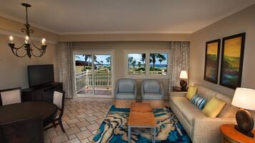 A living room with a TV, dining table, chairs, a sofa, and a view of the beach through large windows.