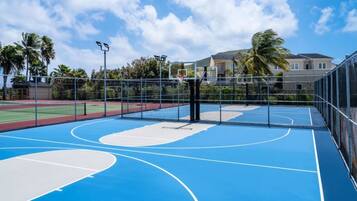 Een buitenbasketbalveld met een blauwe ondergrond, omringd door een metalen hek en palmbomen op de achtergrond.