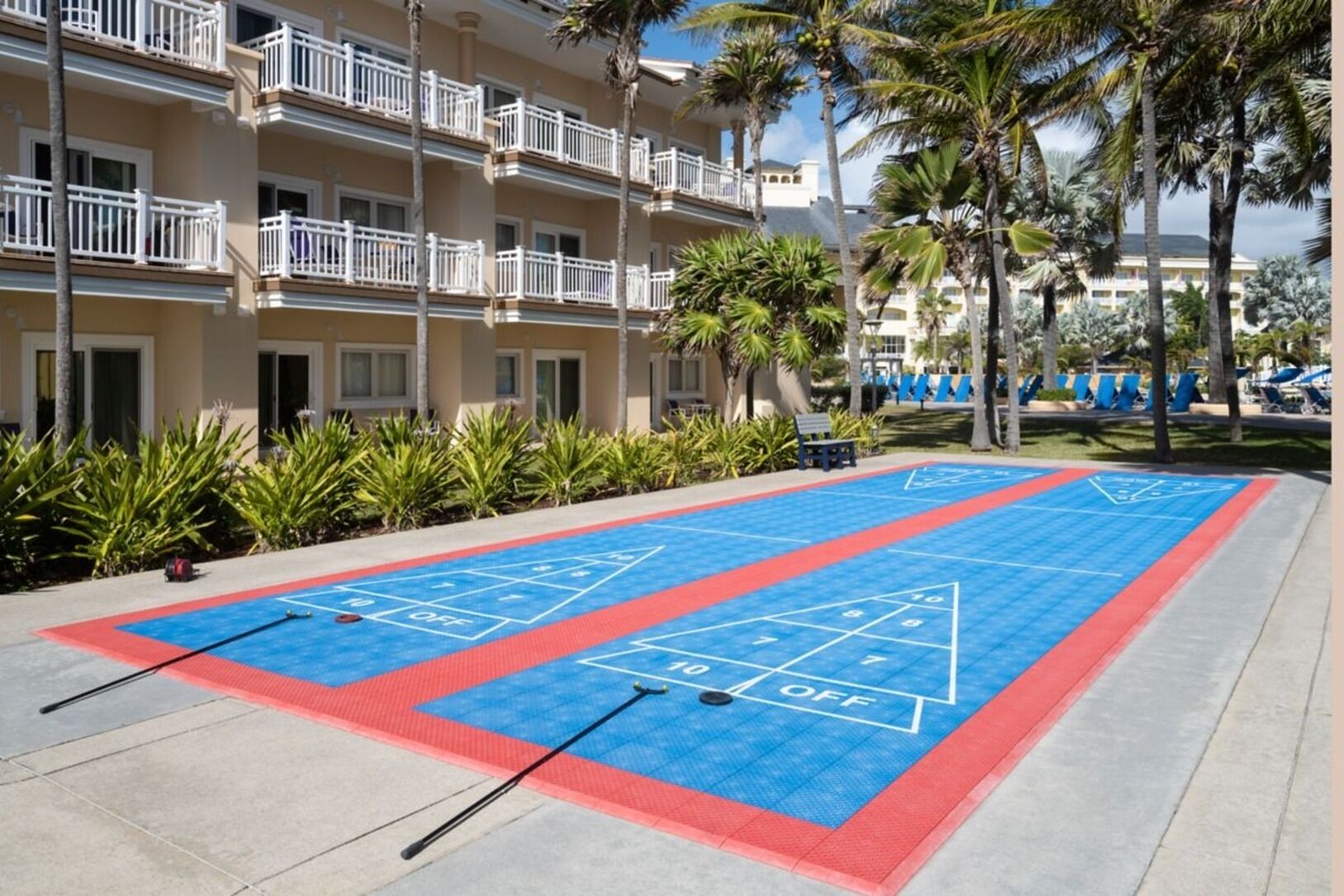 Un terrain de jeu de shuffleboard avec une table de billard, le tout entouré d’un bâtiment doté de balcons et bordé de palmiers.