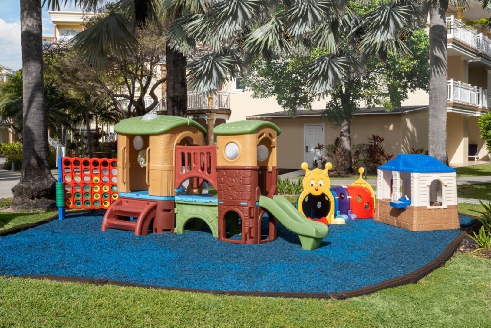 Un terrain de jeux doté de diverses structures ludiques, entouré d’une aire de jeux bleue et de palmiers.