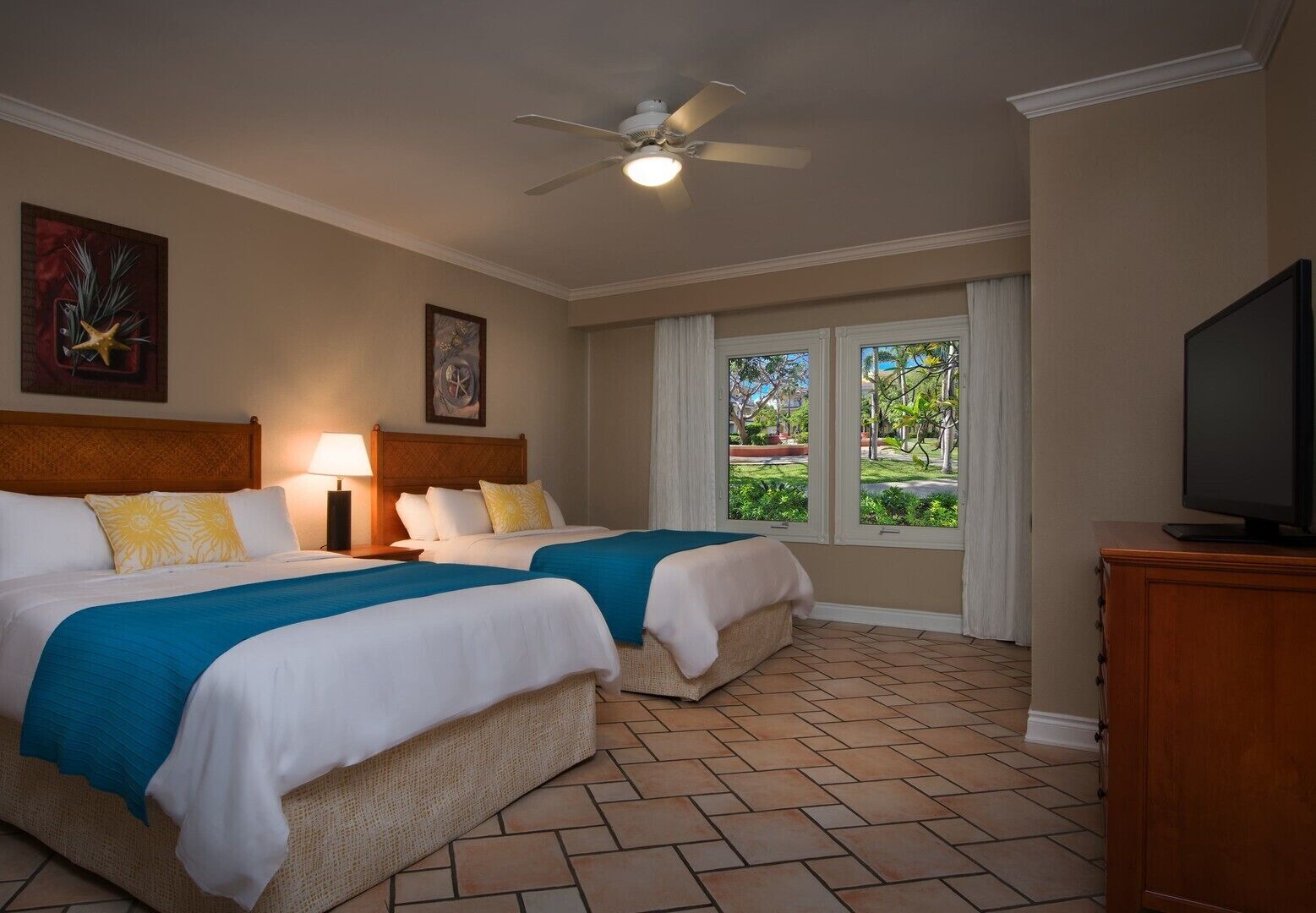 A hotel room with two beds, a ceiling fan, a TV, and a window with a view of greenery.