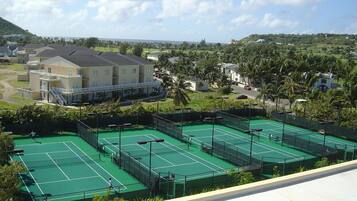 A tennis court complex with multiple courts, a building with balconies, and a view of a residential area with houses and palm trees.