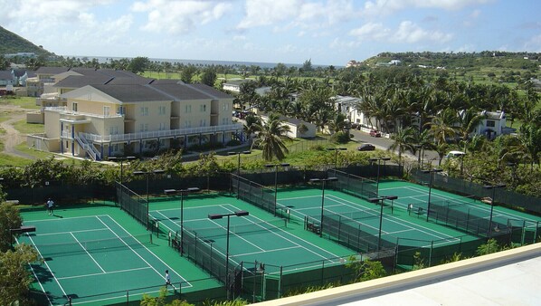 A tennis court complex with multiple courts, a building with balconies, and a view of a residential area with houses and palm trees.