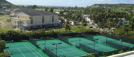 A tennis court complex with multiple courts, a building with balconies, and a view of a residential area with houses and palm trees.