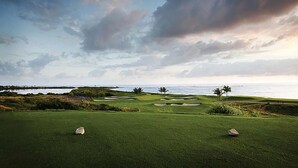 A golf course with bunkers and palm trees, overlooking the ocean.