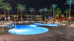 Ein Poolbereich bei Nacht mit Liegestühlen und Sonnenschirmen, umgeben von Palmen und einem Gebäude mit Balkonen.