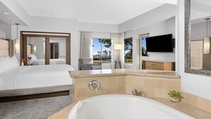 Ein Hotelzimmer mit einem großen Bett, einer separaten Sitzecke mit Sessel und Tisch, einer Badewanne, einem Fernseher und Blick auf Palmen.