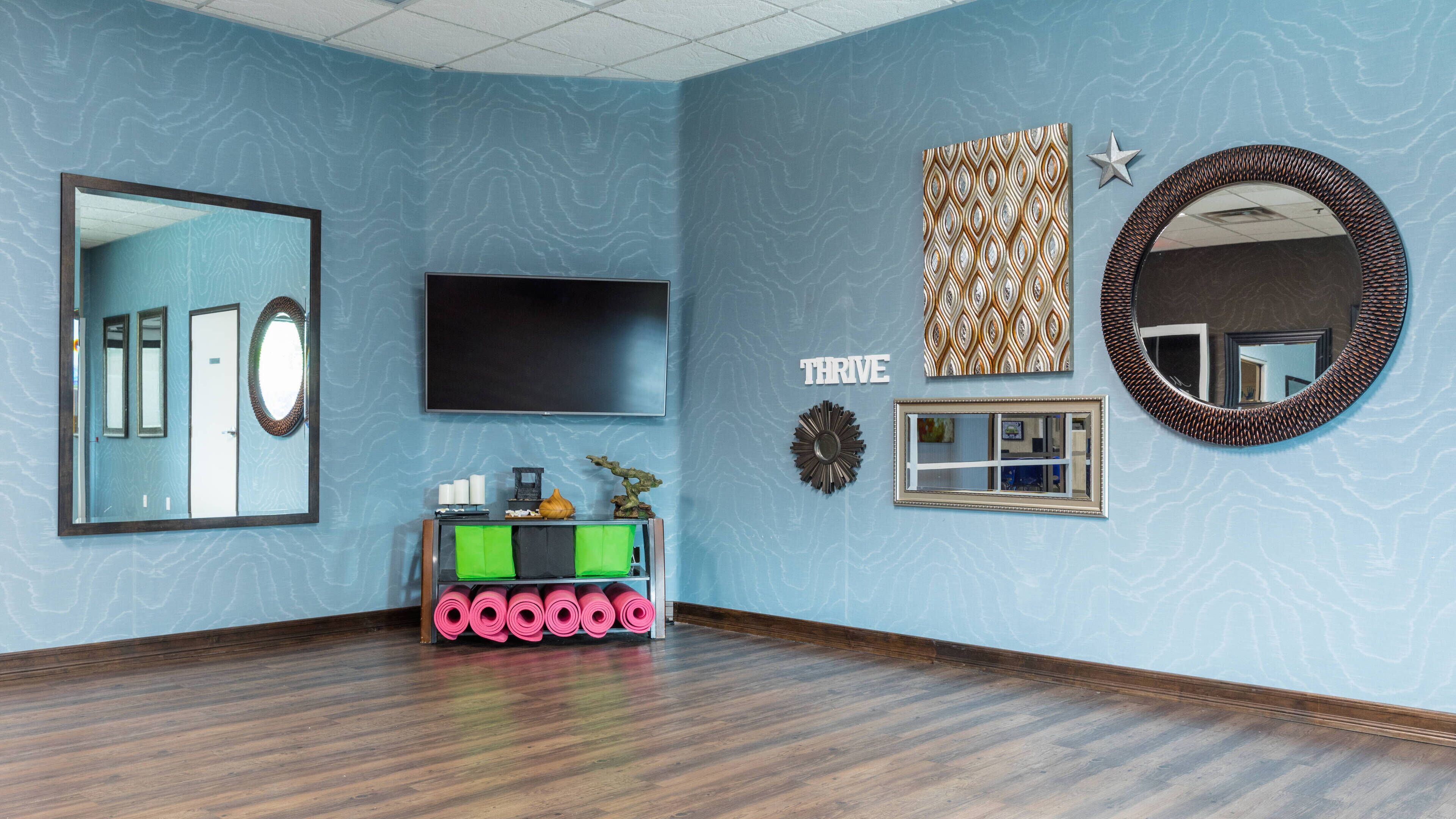 Una habitación con un espejo grande, un televisor de pantalla plana, un espacio para guardar colchoneta de yoga y una obra de arte en la pared con la palabra 'THRIVE'.