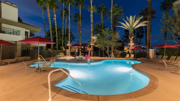 A swimming pool with lounge chairs and umbrellas, surrounded by tall palm trees and a building in the background.