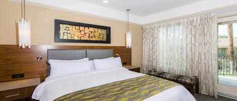 A hotel room with a large bed, a headboard with a geometric pattern, a bedside table, a bench, and a view of the outside through a window with curtains.