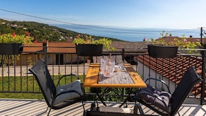 Een balkon met een houten tafel en stoelen, plantenbakken en uitzicht op een kustlandschap.