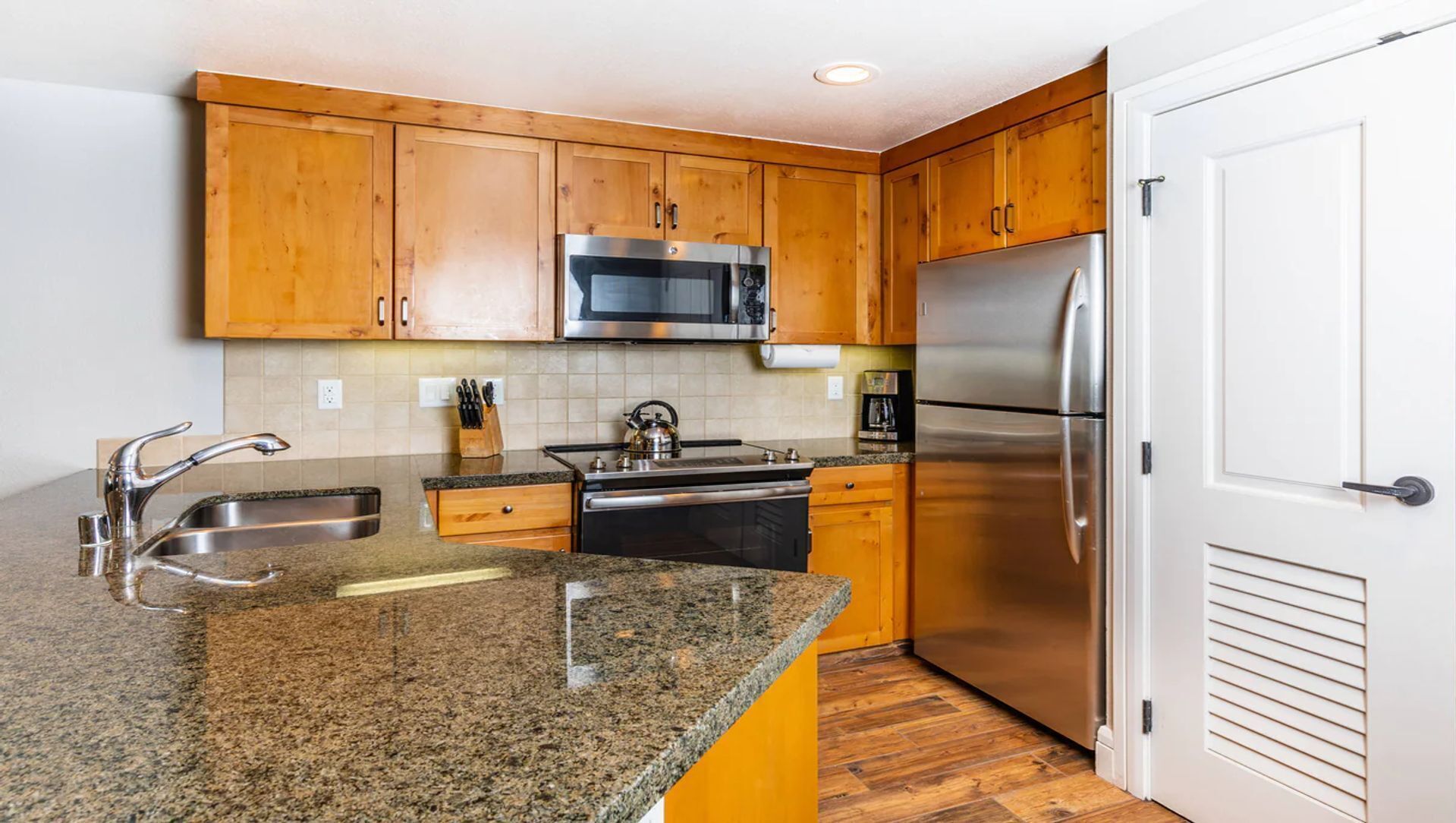 A kitchen with granite countertops, stainless steel appliances, wooden cabinets, and a white door.