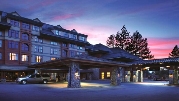 A multi-story hotel with a covered entrance and stone pillars, featuring a large parking area and a view of trees.