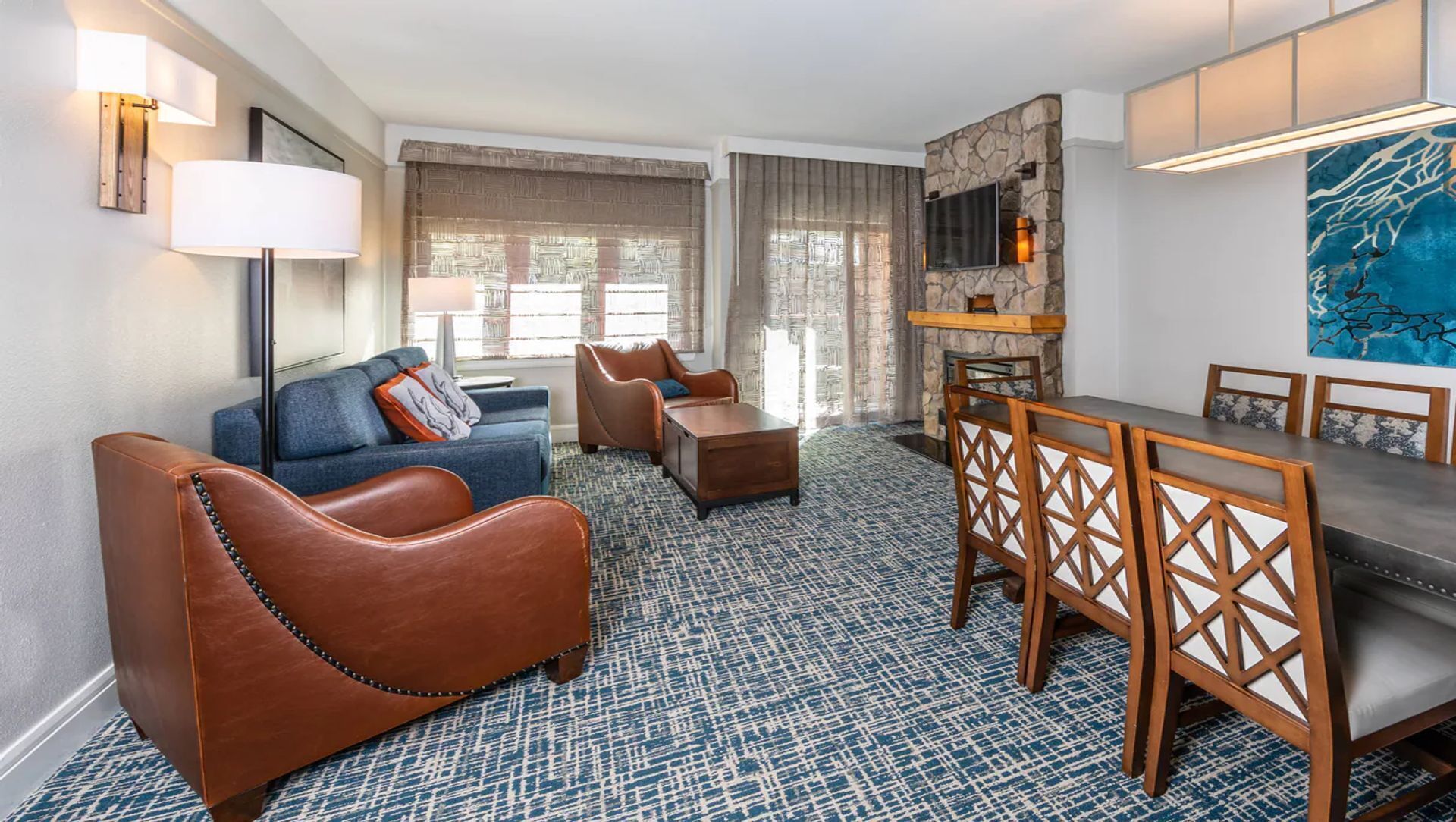 Une chambre d’hôtel moderne avec une moquette à motifs bleus, des fauteuils en cuir, un canapé, une table à manger avec des chaises en bois, un téléviseur à écran plat fixé à un mur en pierre et un grand tableau abstrait.