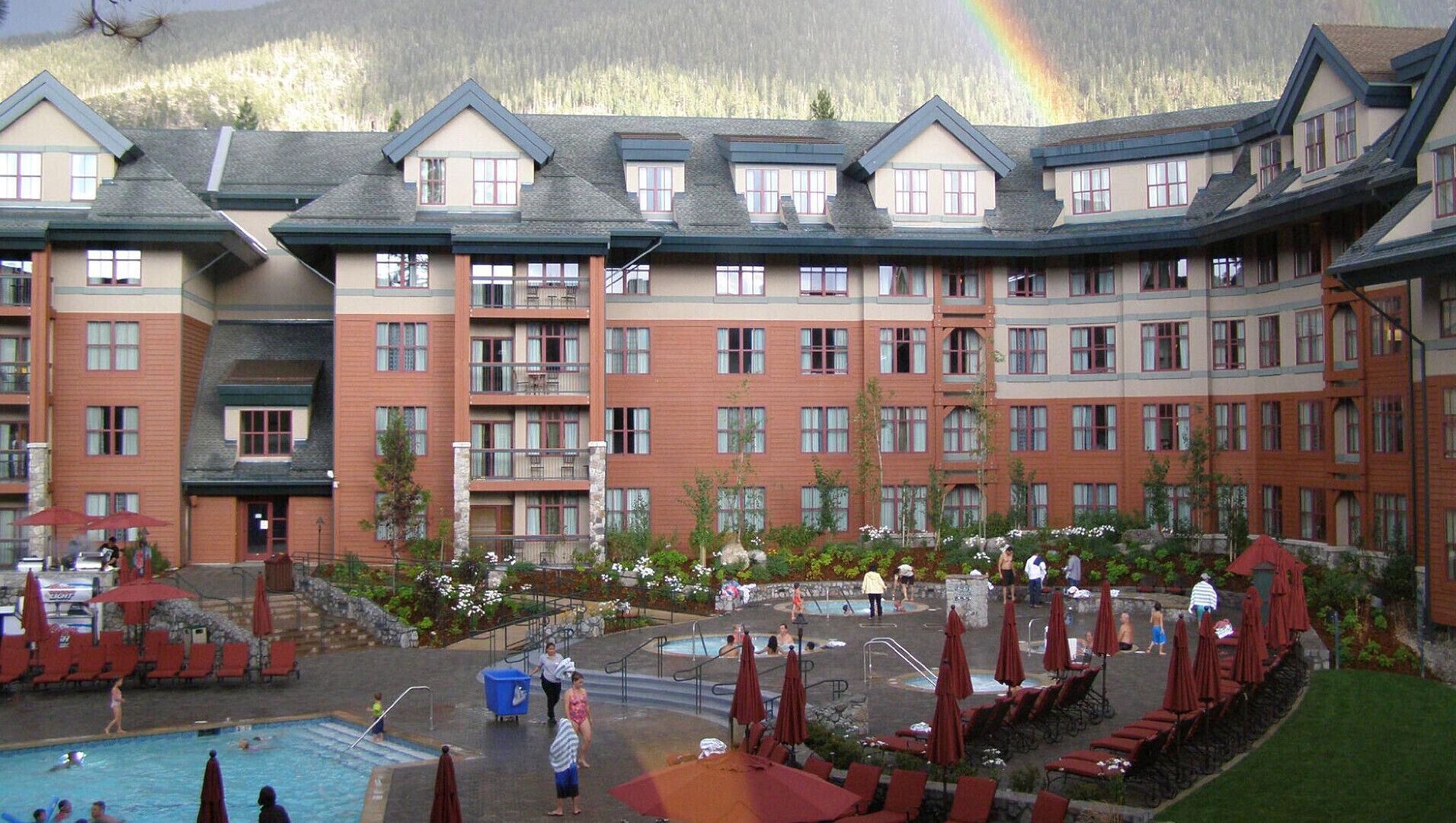 Un hotel con varios pisos, piscina al aire libre y zona de descanso con sombrillas.