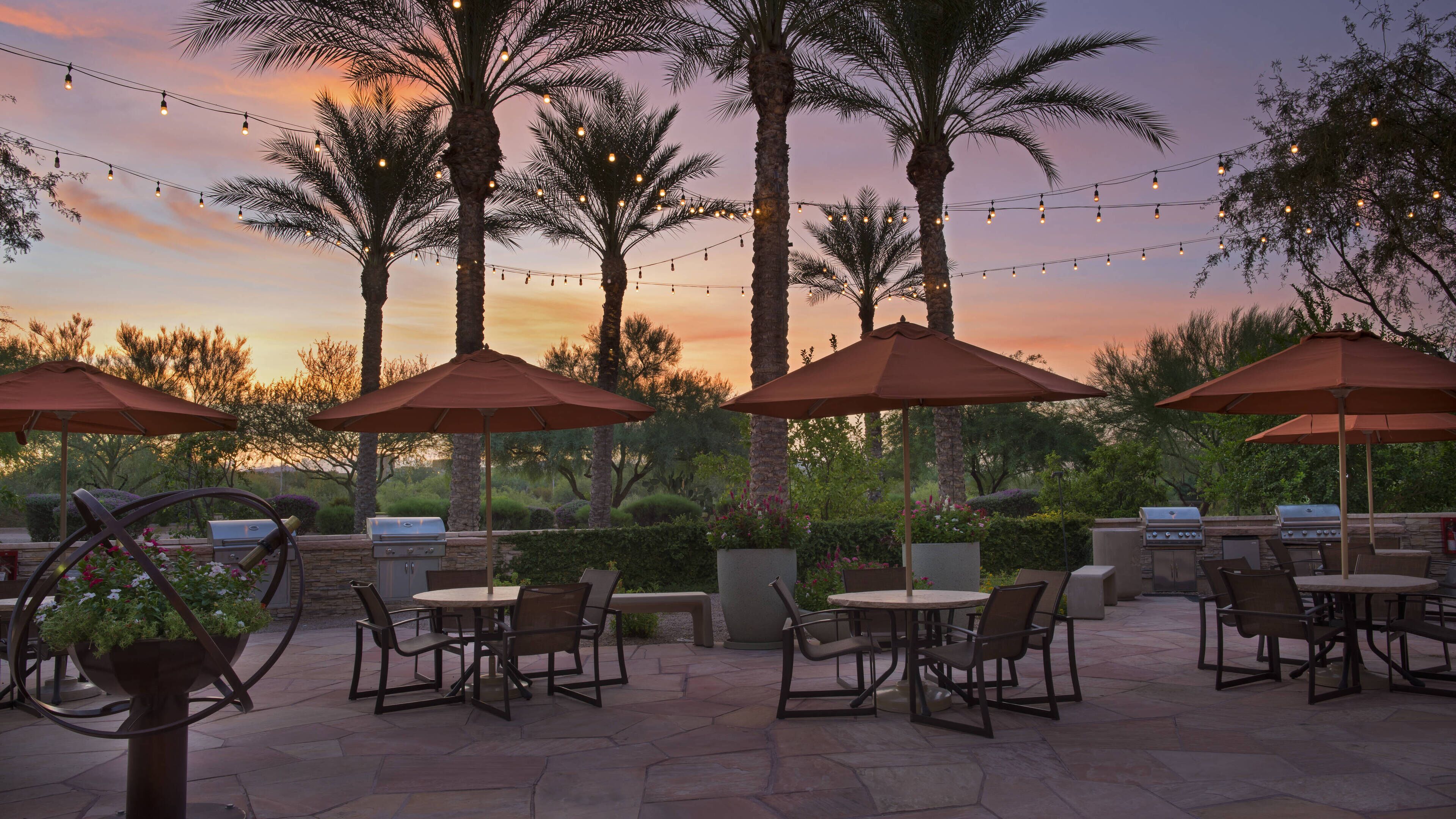 An outdoor patio with stone flooring, multiple barbecue grills, and outdoor seating with umbrellas.