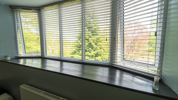 Une pièce dotée d’une grande fenêtre en saillie, agrémentée de stores horizontaux, avec un radiateur en dessous, et offrant une vue sur la verdure environnante.