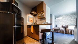 Cocina compacta con refrigerador, microondas y armarios de madera. Hay una pequeña barra con taburetes y una cama en la habitación contigua.