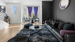 A modern living room with a leather sofa, a round coffee table, and a large mirror. There is a staircase leading to another level, and a view of the outside through large windows with purple curtains. - Stylish Nashville Stay • Skyline Views (Nashville)