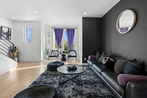 Ein modernes Wohnzimmer mit einer Ledersofa, einem runden Couchtisch und einem großen Spiegel. Eine Treppe führt zu einer weiteren Ebene, und durch große Fenster mit violetten Vorhängen bietet sich ein Blick ins Freie.