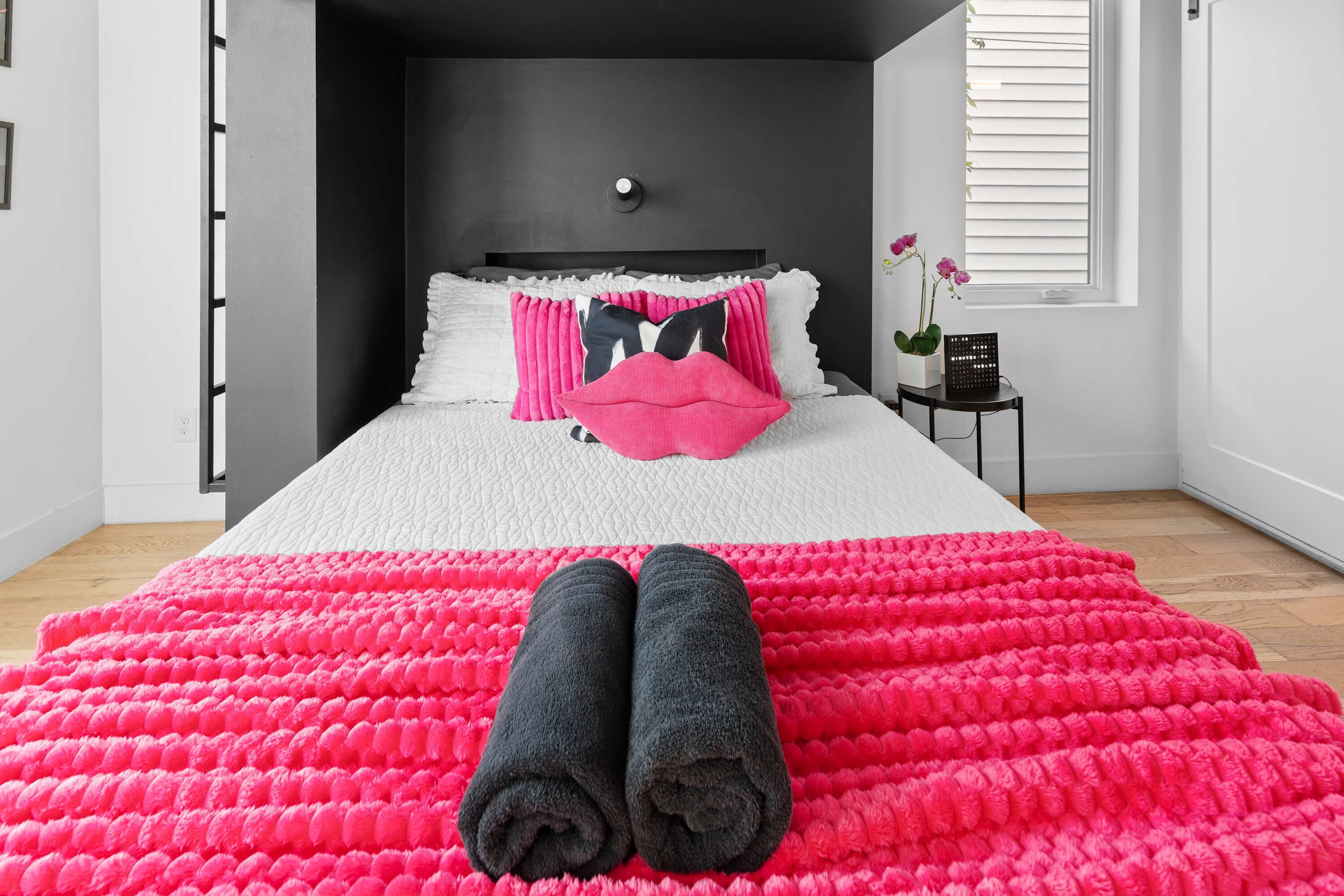 A neatly made bed with a white and pink pillow set, a black and pink pillow, and two rolled towels on a bright pink blanket.