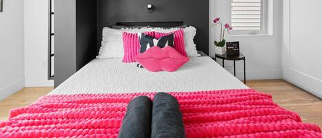 A neatly made bed with a white and pink pillow set, a black and pink pillow, and two rolled towels on a bright pink blanket.