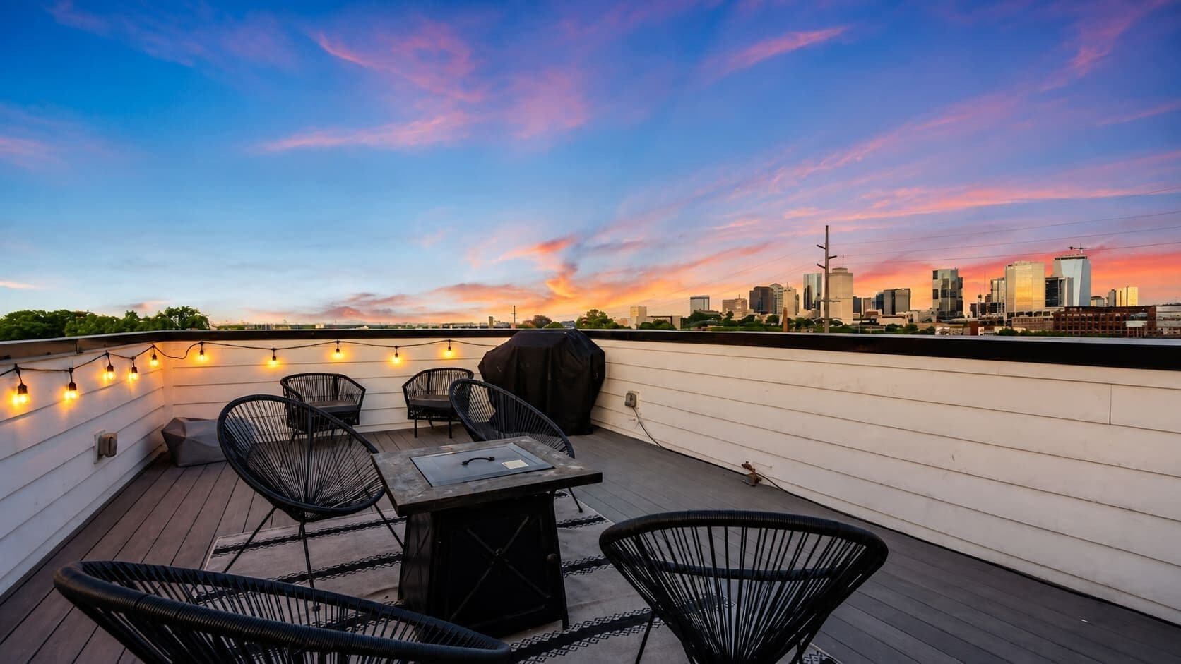 Terraza en la azotea con vistas a un horizonte urbano, mobiliario exterior e iluminación con luces colgantes.