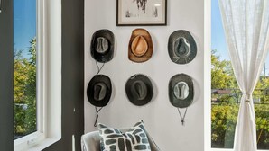 Una habitación con un sillón gris, una colección de sombreros colgada en la pared, un cuadro abstracto enmarcado y vistas a través de una ventana con cortinas blancas.